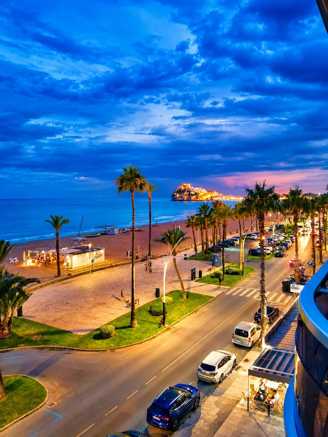 Vue au coucher du soleil depuis l'appartement : avenue, promenade bordée de palmiers, plage et château de Peñíscola